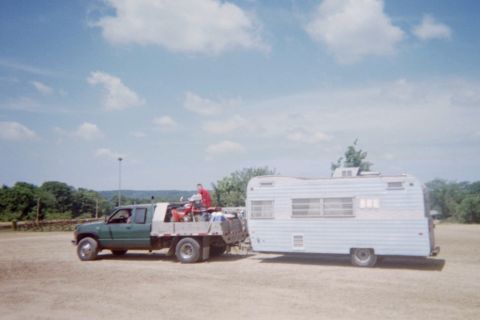 1976 Jeep CJ5 - A green flatbed truck tows a white camper trailer across a dusty lot under a partly cloudy sky.