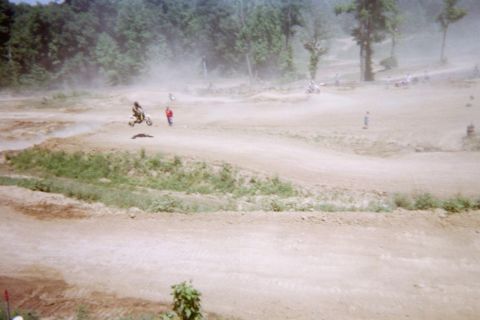 1976 Jeep CJ5 - Motocross riders navigate a dusty dirt track with jumps and turns, surrounded by trees.