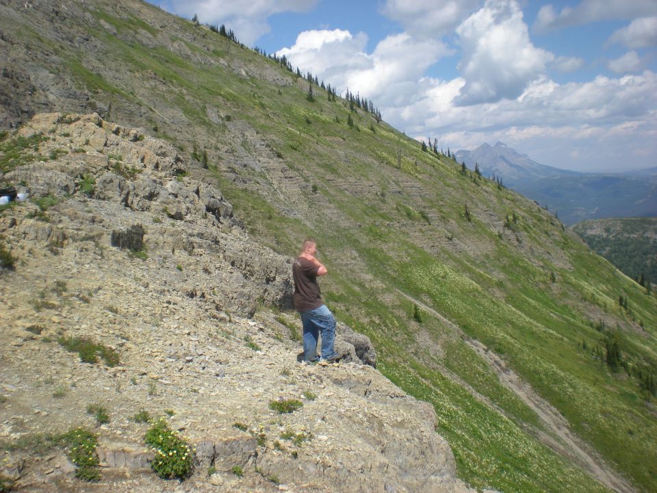 Man in brown shirt and jeans stands on rocky outcrop overlooking a grassy mountain slope with scattered trees under a cloudy sky