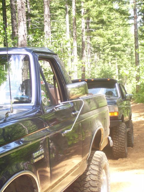 1991 Ford Bronco - Two dark SUVs, including a 1991 Ford Bronco, are parked side-by-side on a dirt road surrounded by trees.