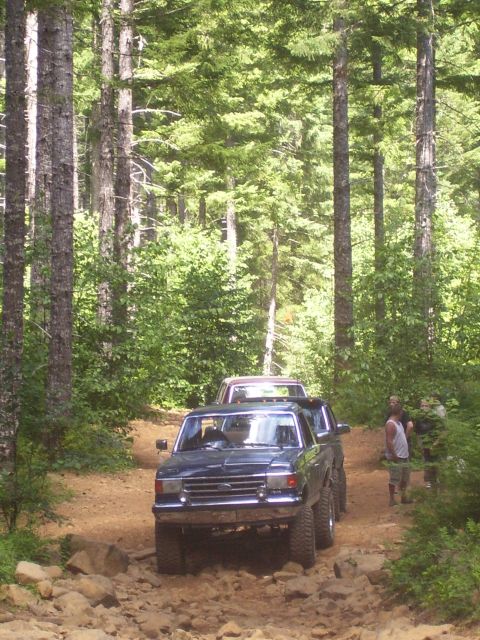 1991 Ford Bronco - Two trucks navigate a rough, forested path, with people standing by the side of the road.
