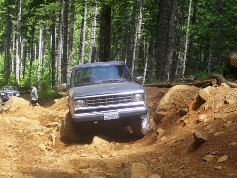1991 Ford Bronco - A Ford Ranger tackles a challenging off-road course in a wooded area, with another vehicle following.