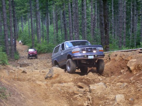 1991 Ford Bronco - A lifted blue Ford Bronco navigates a challenging off-road trail with a red Jeep in the background.