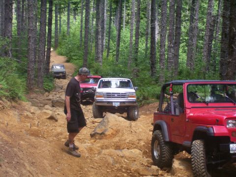 1991 Ford Bronco - Multiple off-road vehicles navigate a challenging, rocky path through a dense forest.