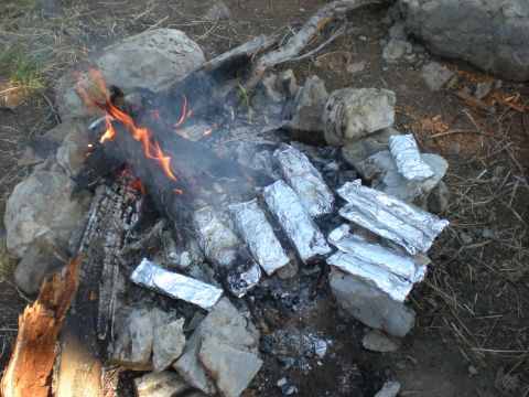 1974 Ford Ranchero - Several foil-wrapped food items are being cooked over a campfire, surrounded by rocks and charred wood.