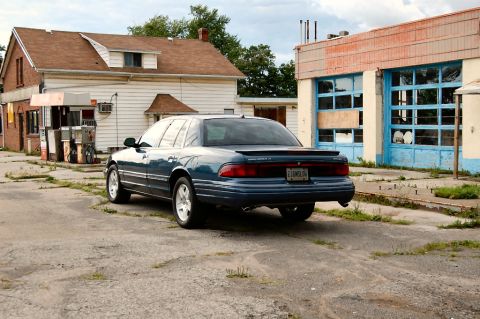 1985 Mercury Grand Marquis - A blue sedan is parked in front of a derelict building, its rear end facing the camera.