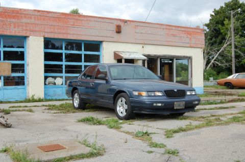 1985 Mercury Grand Marquis - Blue sedan parked in front of an abandoned building with an orange car in the background.