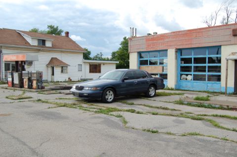 1985 Mercury Grand Marquis - A sedan sits in the cracked asphalt lot of a disused service station under a cloudy sky.