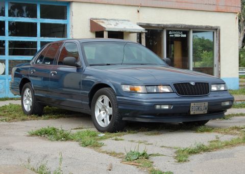 1985 Mercury Grand Marquis - This dark blue sedan, likely a Mercury Grand Marquis from the late 1990s, is parked in front of a building with a garage door.