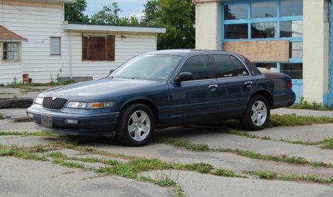 1985 Mercury Grand Marquis - Blue sedan parked on cracked concrete in front of a building with a garage door.