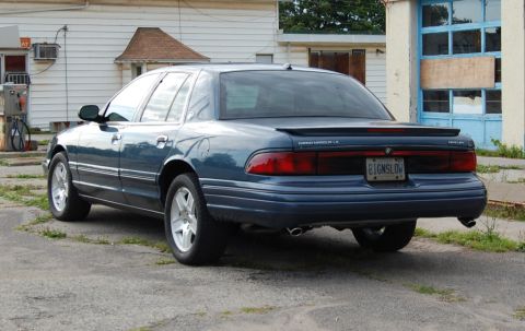 1985 Mercury Grand Marquis - Rear view of a blue sedan with a spoiler and custom license plate parked in front of a building.