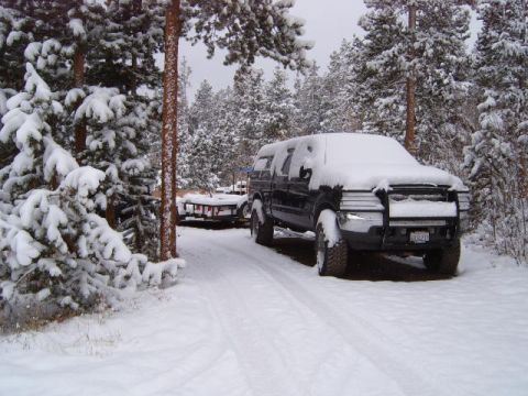 1956 Ford Fairlane - A black pickup truck sits covered in snow on a winter day, parked on a snowy road surrounded by pine trees.