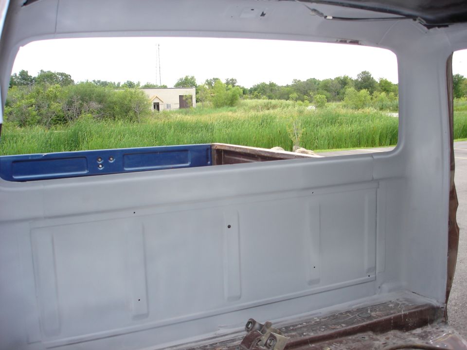 Gray truck bed interior with blue tailgate, looking out at a field and building
