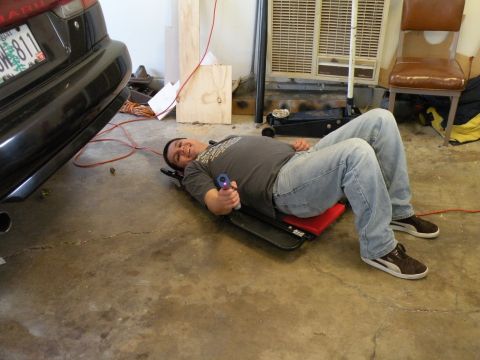 1998 Subaru Legacy - A person lies on a creeper beneath a car, holding a tool, in what appears to be a garage.