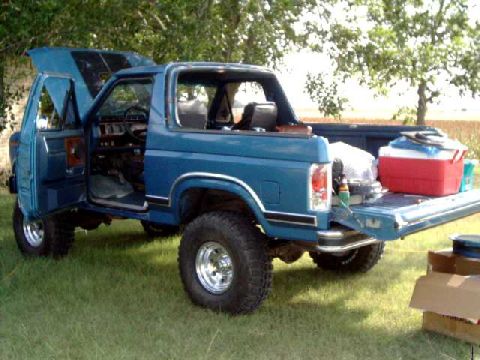 1984 Ford Bronco - 1984 Ford Bronco parked on grass with its hood and driver's door open, tailgate lowered.