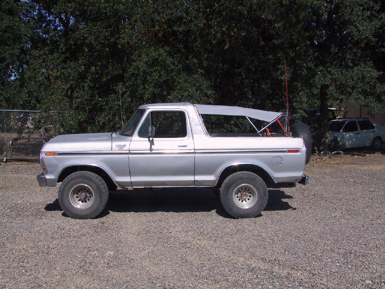 Silver Ford Bronco with a soft top, parked on gravel with trees behind it. Another car is visible in the background
