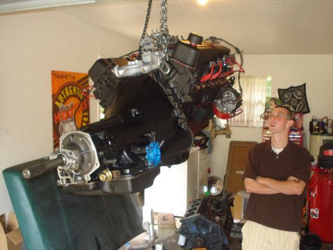 1978 Ford Bronco - A person looks up at a large engine and transmission assembly suspended by chains in a garage.
