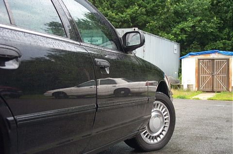 2001 Ford Crown Victoria - A black 2001 Ford Crown Victoria is seen from the side, with a reflection of a white car visible on its wet surface.
