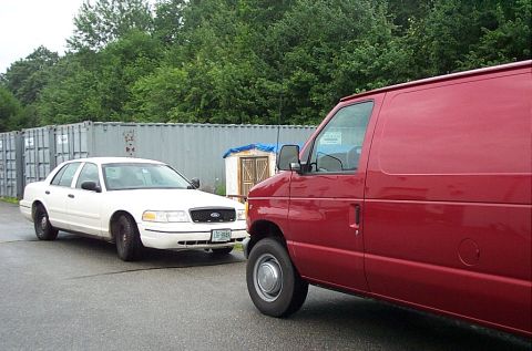 2001 Ford Crown Victoria - A white 2001 Ford Crown Victoria is parked next to a red van in an outdoor lot with shipping containers in the background.