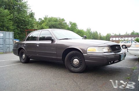 2001 Ford Crown Victoria - A gray Ford Crown Victoria police car is parked in a lot with trees and buildings in the background.