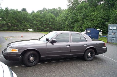 2001 Ford Crown Victoria - A dark gray 2001 Ford Crown Victoria sedan is parked on asphalt with trees in the background.