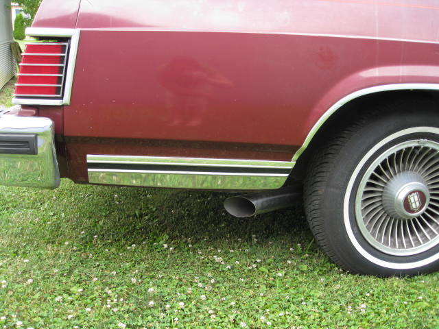 Rear quarter of maroon car with vertical taillight, chrome exhaust tip and wire wheel cover on grass