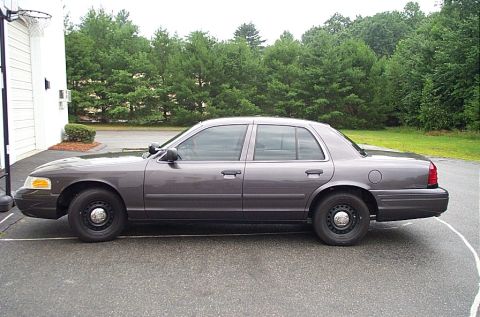 2001 Ford Crown Victoria - A gray 2001 Ford Crown Victoria is parked on an asphalt surface next to a building with trees in the background.