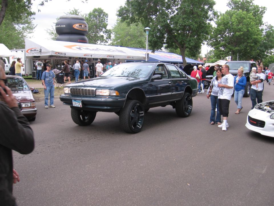 Dark green Chevrolet Caprice on tall lift with black wheels at a car show
