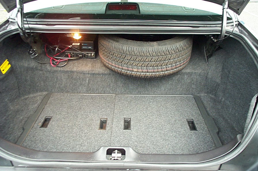 Trunk interior of a 2001 Ford Crown Victoria with a spare tire, a black electronic device with red wires, and gray carpeting