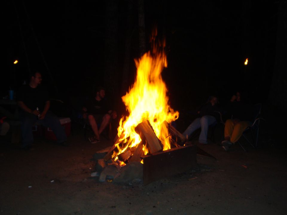 Large nighttime campfire with people seated around it in folding chairs