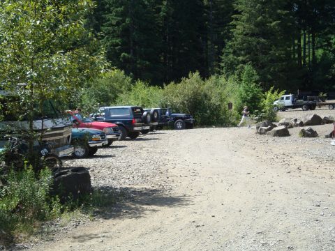 1978 Ford Bronco - A gravel lot is filled with parked vehicles, including SUVs and trucks, with a wooded area in the background.