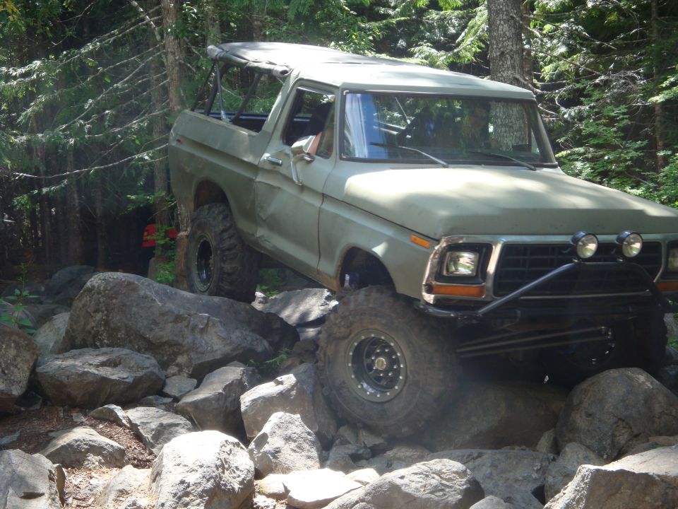 Olive green 1978 Ford Bronco off-roading over large rocks on a wooded trail