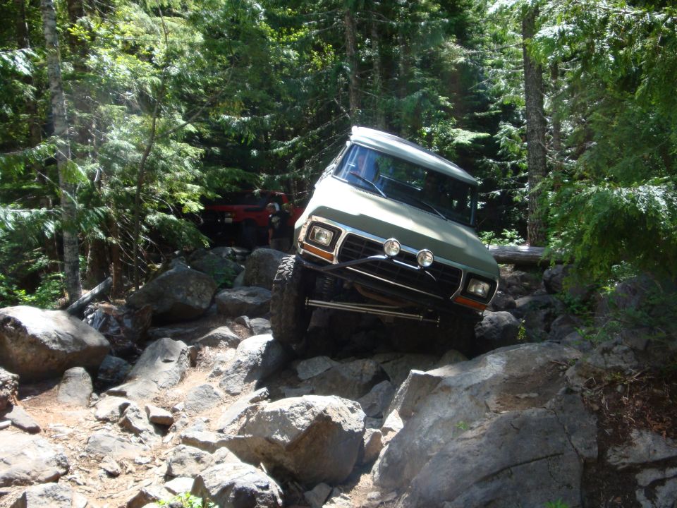 1978 Ford Bronco climbing over large rocks on a wooded trail, another red vehicle behind it