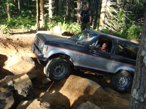 1978 Ford Bronco - A woman drives a silver and black SUV through a challenging rocky off-road course in a wooded area.