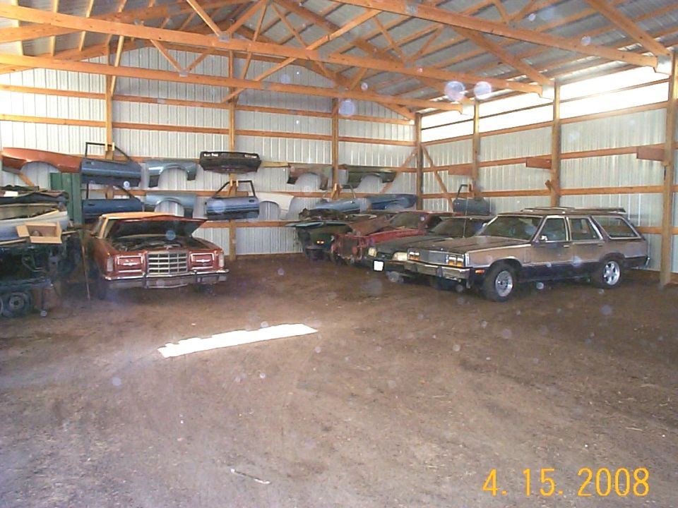Barn interior with multiple vehicles and car parts, including a brown station wagon and a red sedan