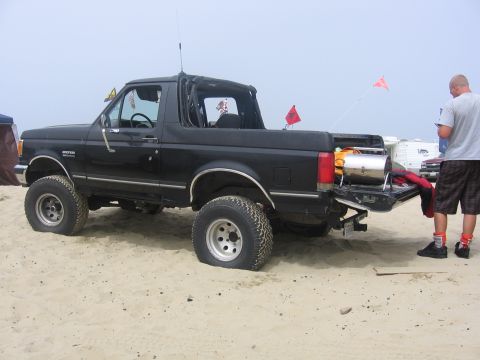 1989 Ford Bronco - This black Ford Bronco is parked on the sand with its tailgate open, and a person is standing nearby.