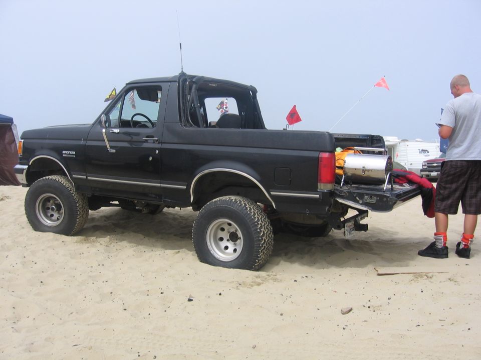 Black Ford Bronco with flat tires on the beach. A person stands by the open tailgate