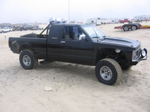1989 Ford Bronco - This black pickup truck, modified for off-road use, sits on a sandy terrain with other vehicles and trailers in the background.