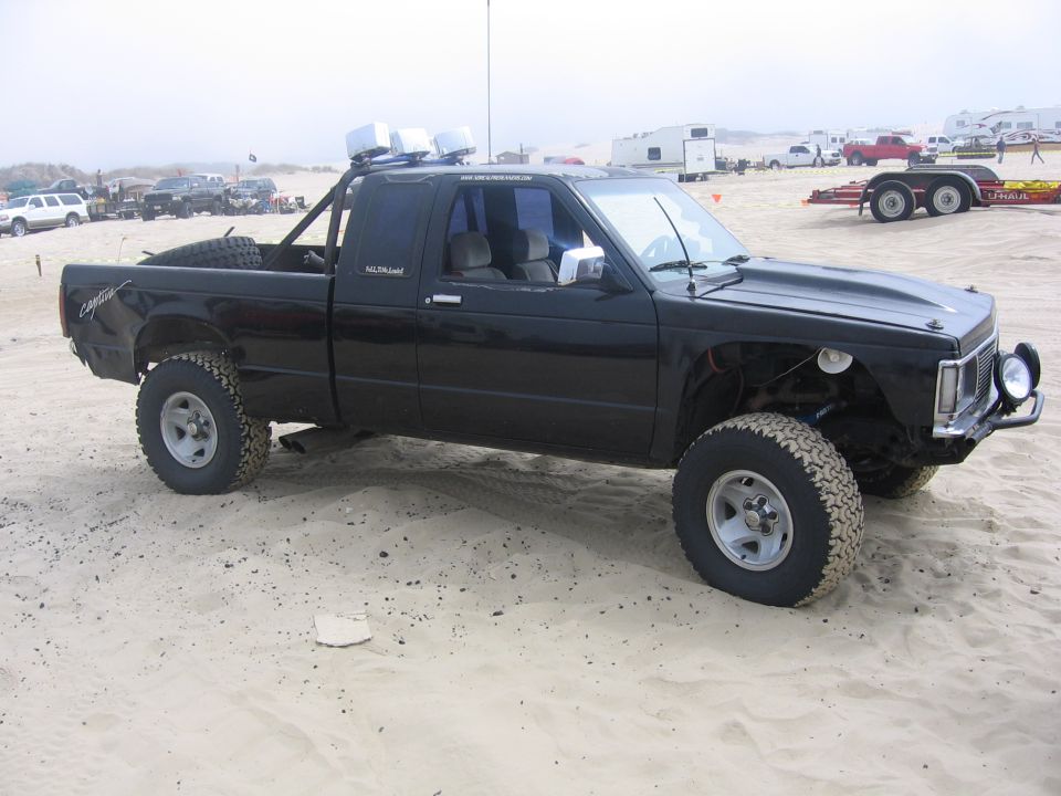 Black pickup truck with off-road tires and light bar on sand, viewed from the front three-quarter angle