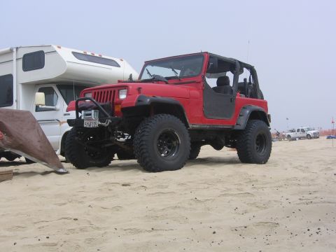 1989 Ford Bronco - A red Jeep Wrangler is parked on a sandy beach next to an RV, with a white pickup truck visible in the background.