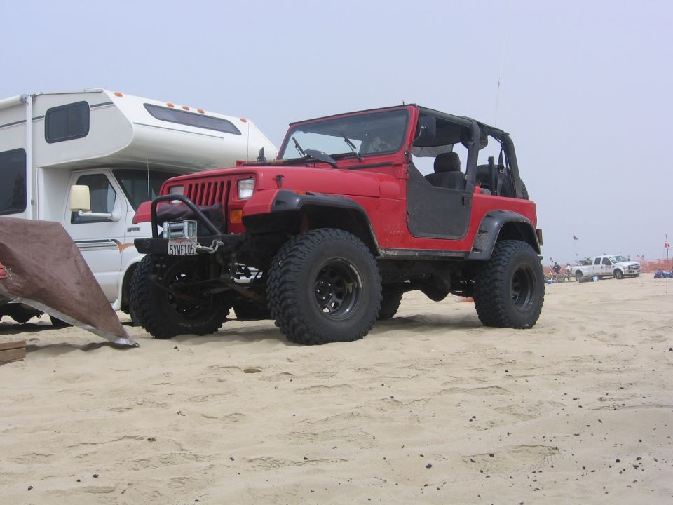 Red Jeep Wrangler with aftermarket bumper and large tires on a sandy beach