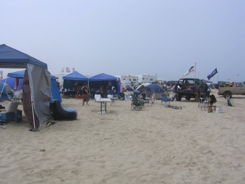 1989 Ford Bronco - A busy beach campsite with multiple tents and vehicles set up on the sand under an overcast sky.