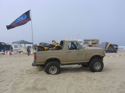 1989 Ford Bronco - A tan 1989 Ford Bronco is parked on the sand, carrying a yellow ATV in its bed, with tents and other vehicles in the background.