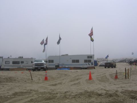 1989 Ford Bronco - A white pickup truck is parked on a sandy beach, surrounded by RVs and various flags under a hazy sky.