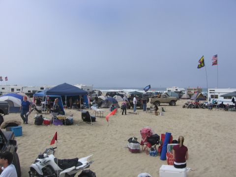 1989 Ford Bronco - A busy beach campsite scene with multiple vehicles, tents, and RVs set up on the sand.