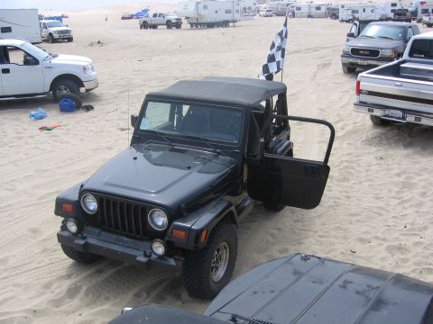 1989 Ford Bronco - A black Jeep Wrangler sits on a sandy beach with its door open, a checkered flag waving from its roll cage.