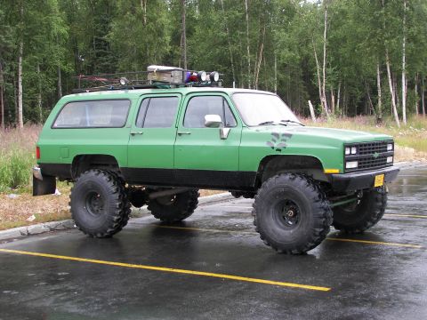 1990 Chevrolet Suburban - Green square-body Chevrolet Suburban lifted on huge military-style bogger tires and black steel wheels, parked in a wet lot backed by birch…