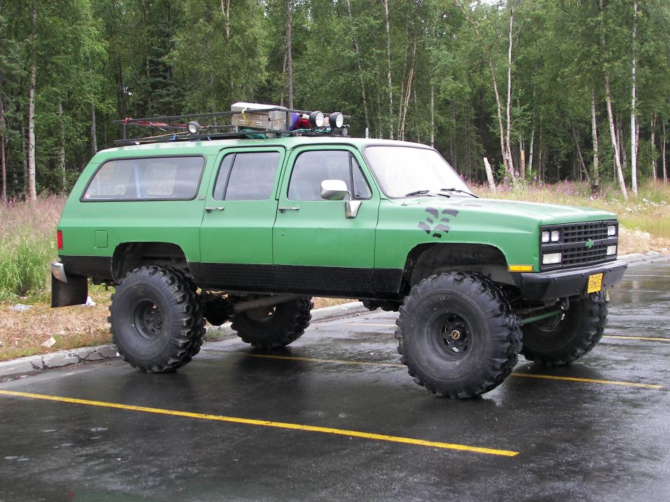 Green lifted Chevrolet Suburban on oversized military tires with roof rack and light bar