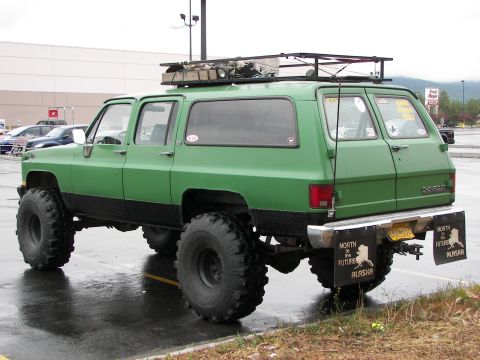 1990 Chevrolet Suburban - This lifted green SUV features oversized tires, a roof rack, and mud flaps with "North to the Future Alaska" text.