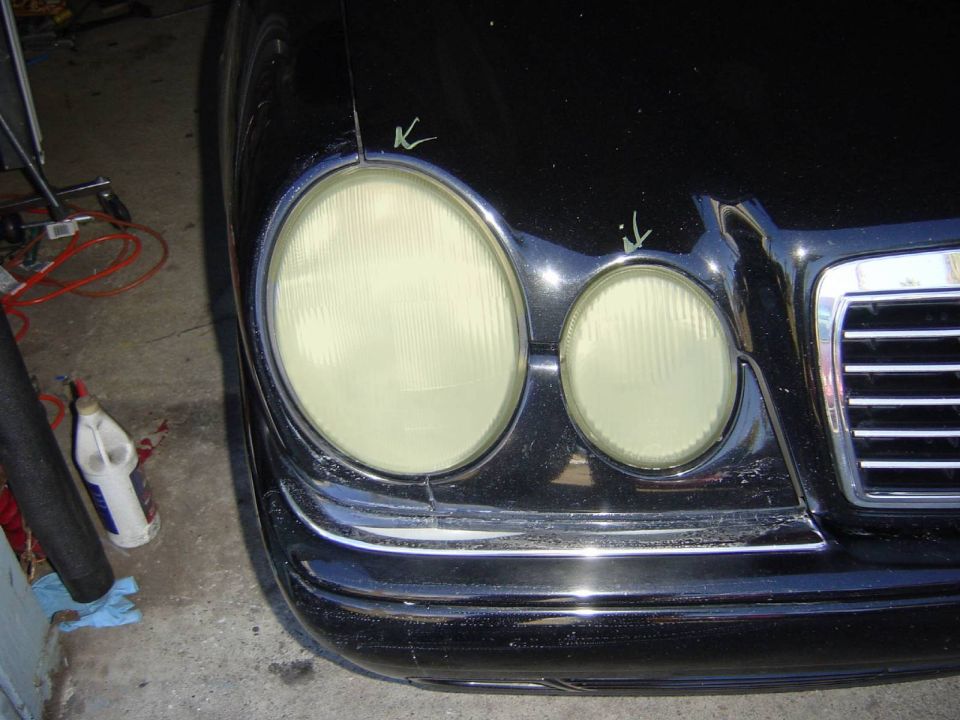 Front corner of a black Mercedes-Benz E-Class (W210) showing the headlights and grille. Green marks are visible on the hood near the headlights